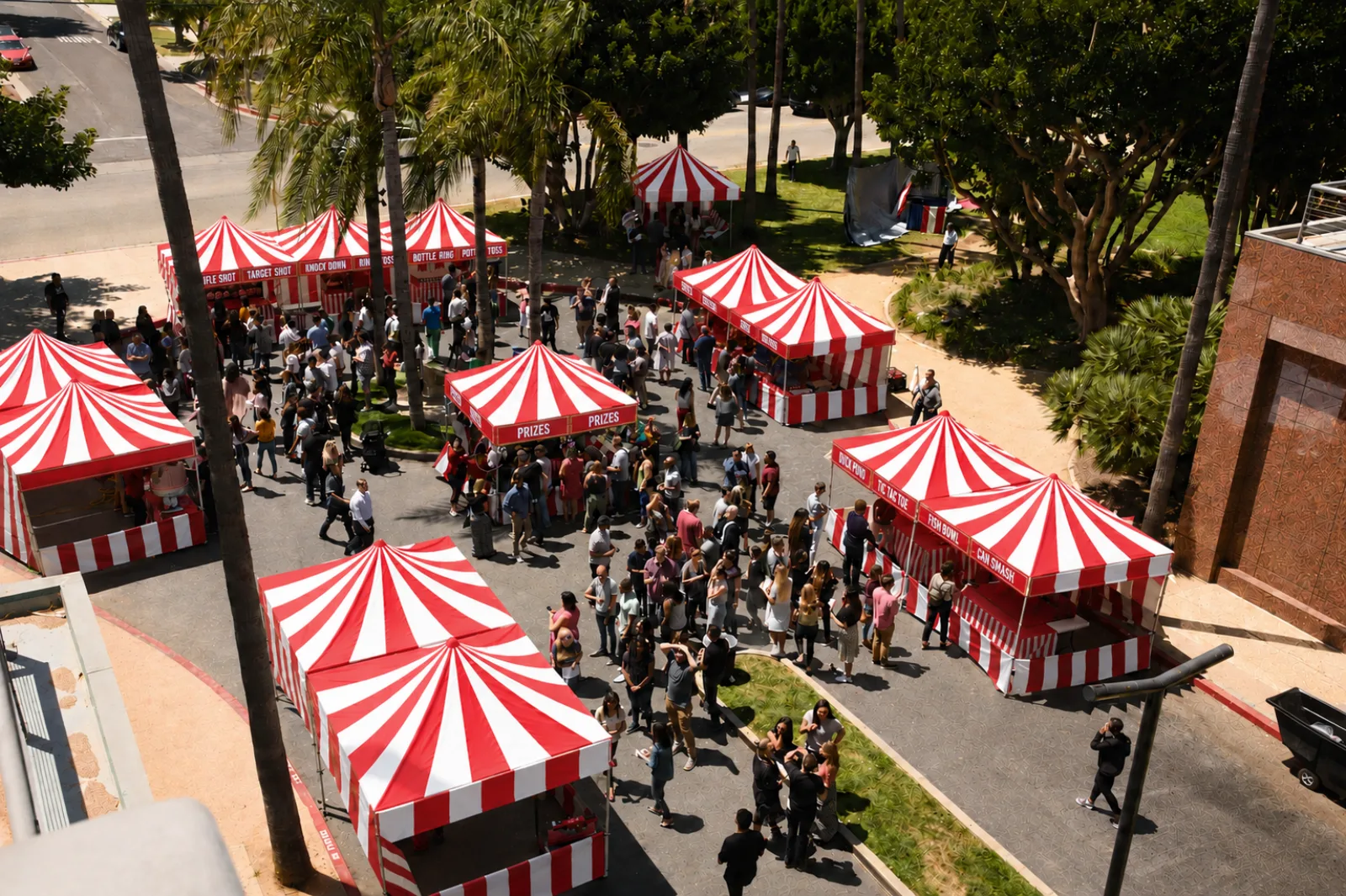 A large public event seen from above, red-and-white striped carnival booths lining a paved courtyard while guests queue at the game counters in Rainbow, CA