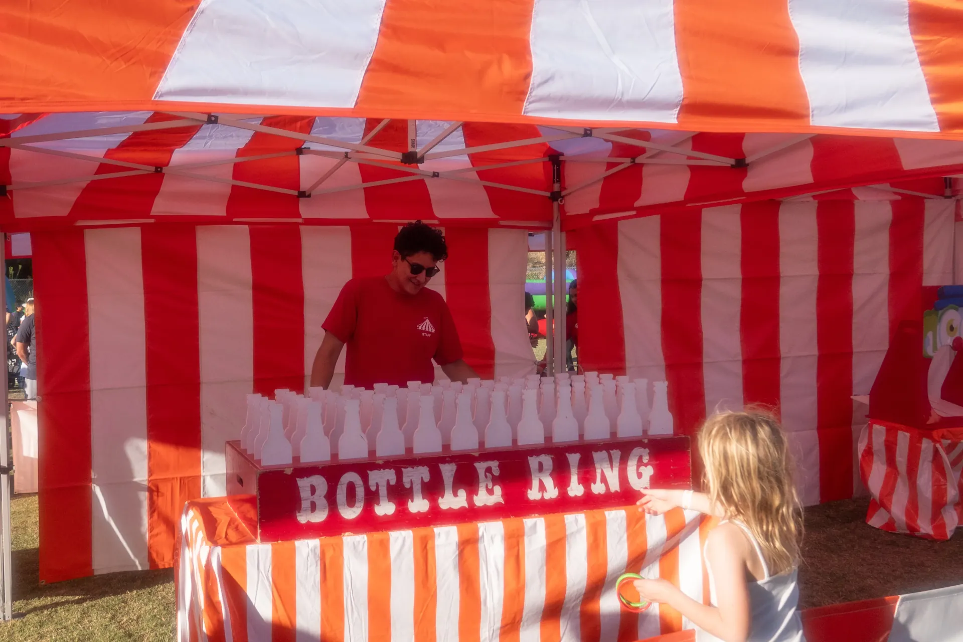 A row of striped carnival game booths with ring toss, bottle ring, and balloon pop set up on a lawn, prizes hanging visibly from each booth in Poway, CA