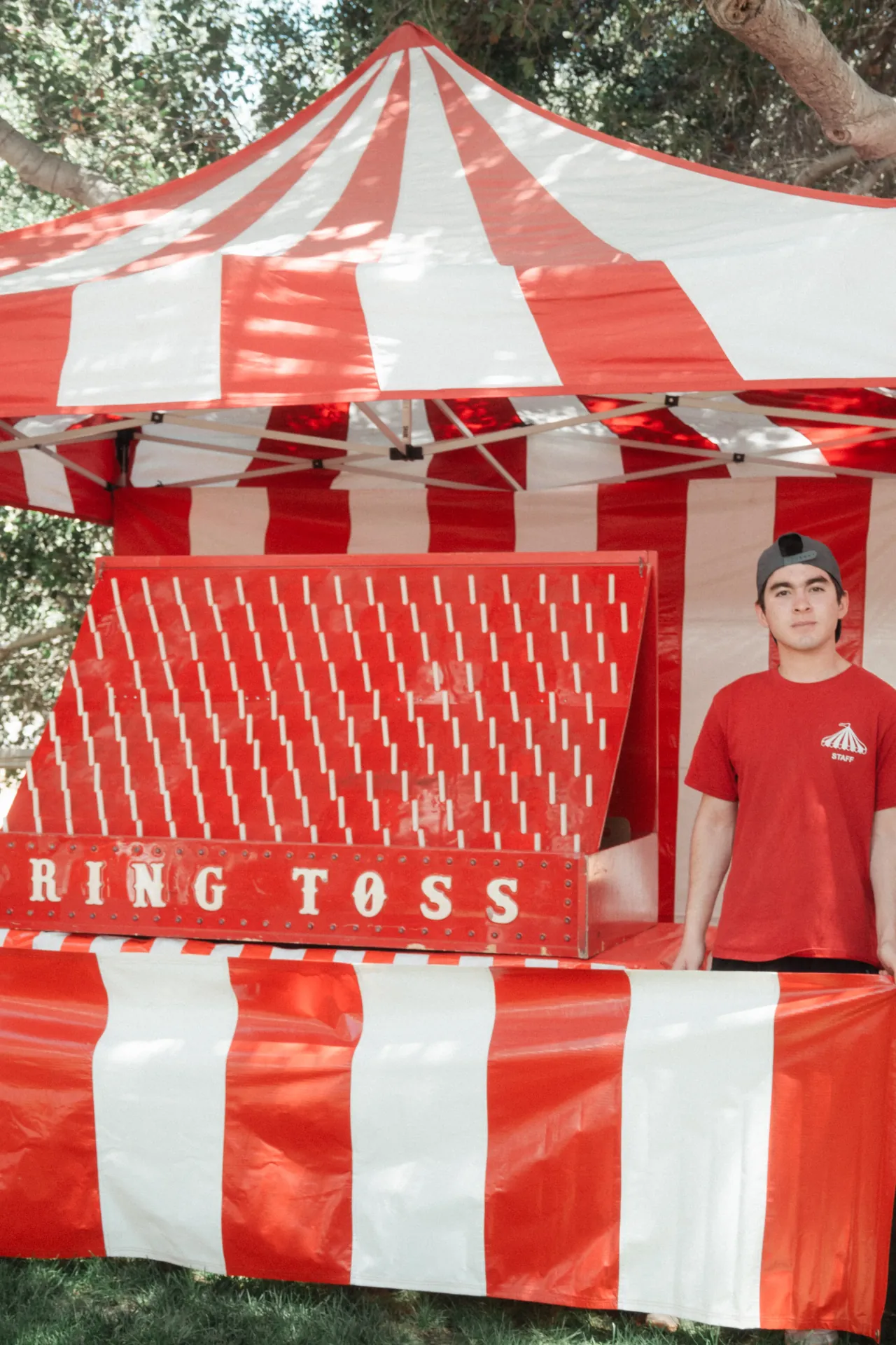 Kids playing a ring toss carnival game booth at an outdoor event, prizes displayed behind the attendant