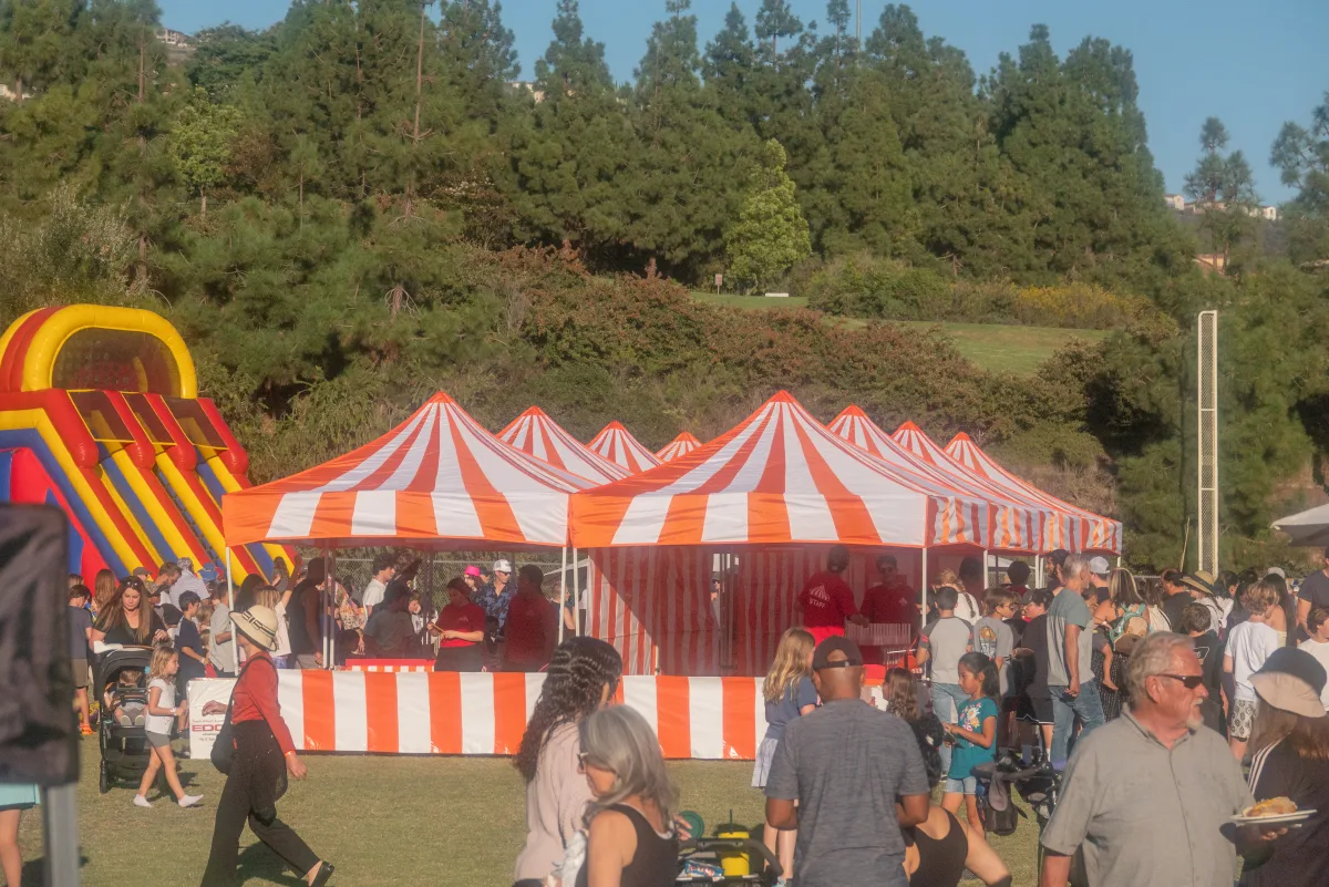 A row of orange-and-white striped carnival booths on a grass field at a busy outdoor event, a tall red-and-yellow inflatable slide at the left edge, and a crowd of families moving through in late-afternoon sun with a wooded hillside behind, one of the three Halloween booth colors available for Rainbow