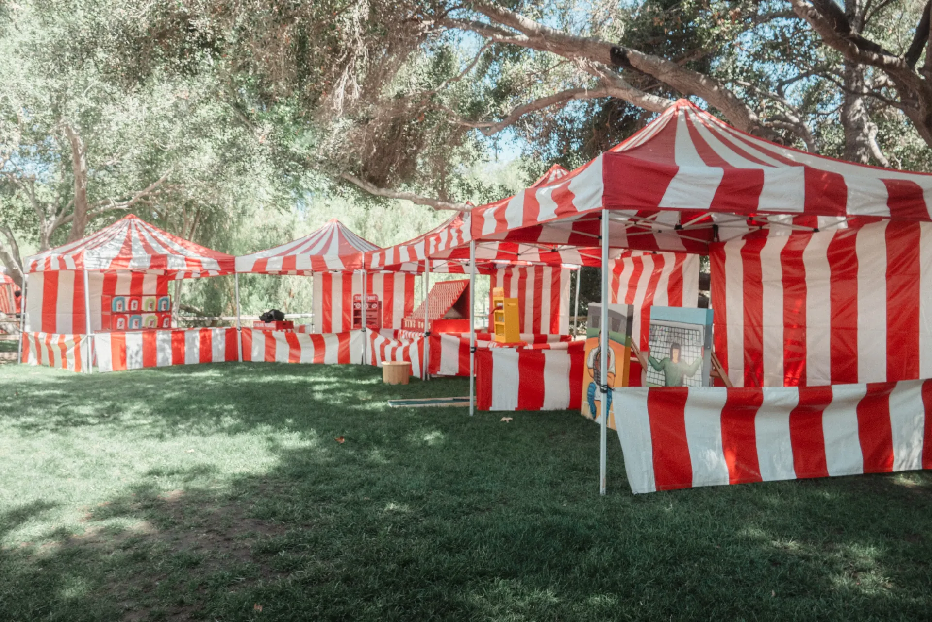 Attendants in striped vests running a row of carnival games at an outdoor community event with a crowd of families moving through a wide midway