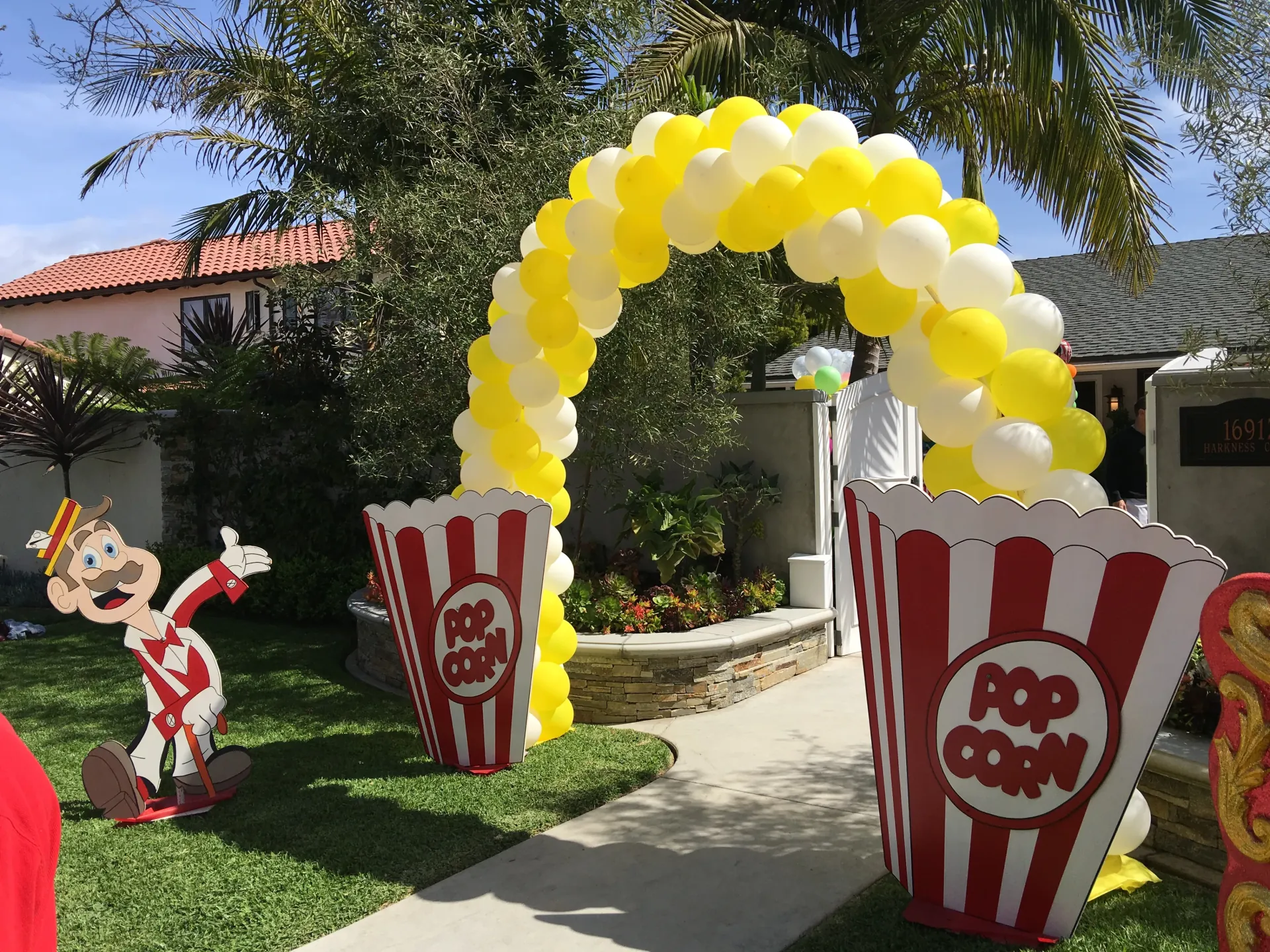 A yellow-and-white balloon arch over a walkway, anchored at each end by a large red-and-white striped popcorn-box prop, with a flat cutout of a moustached carnival barker figure standing on the grass at left, residential landscaping and a tile roof behind