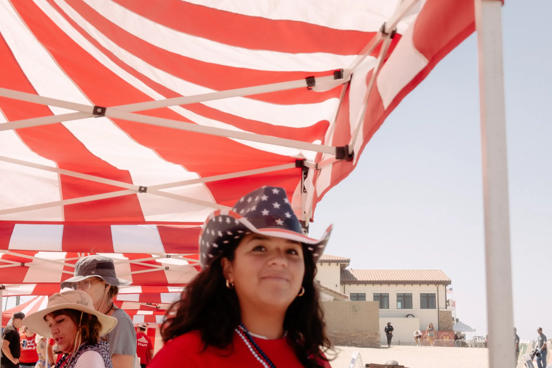 A close view up into the underside of a red-and-white striped canopy on a sunny beach, with a guest in a stars-and-stripes cowboy hat and patriotic bead necklaces in the foreground, other guests in sun hats beside her, and a tile-roofed building on the sand behind in Camp Pendleton, CA