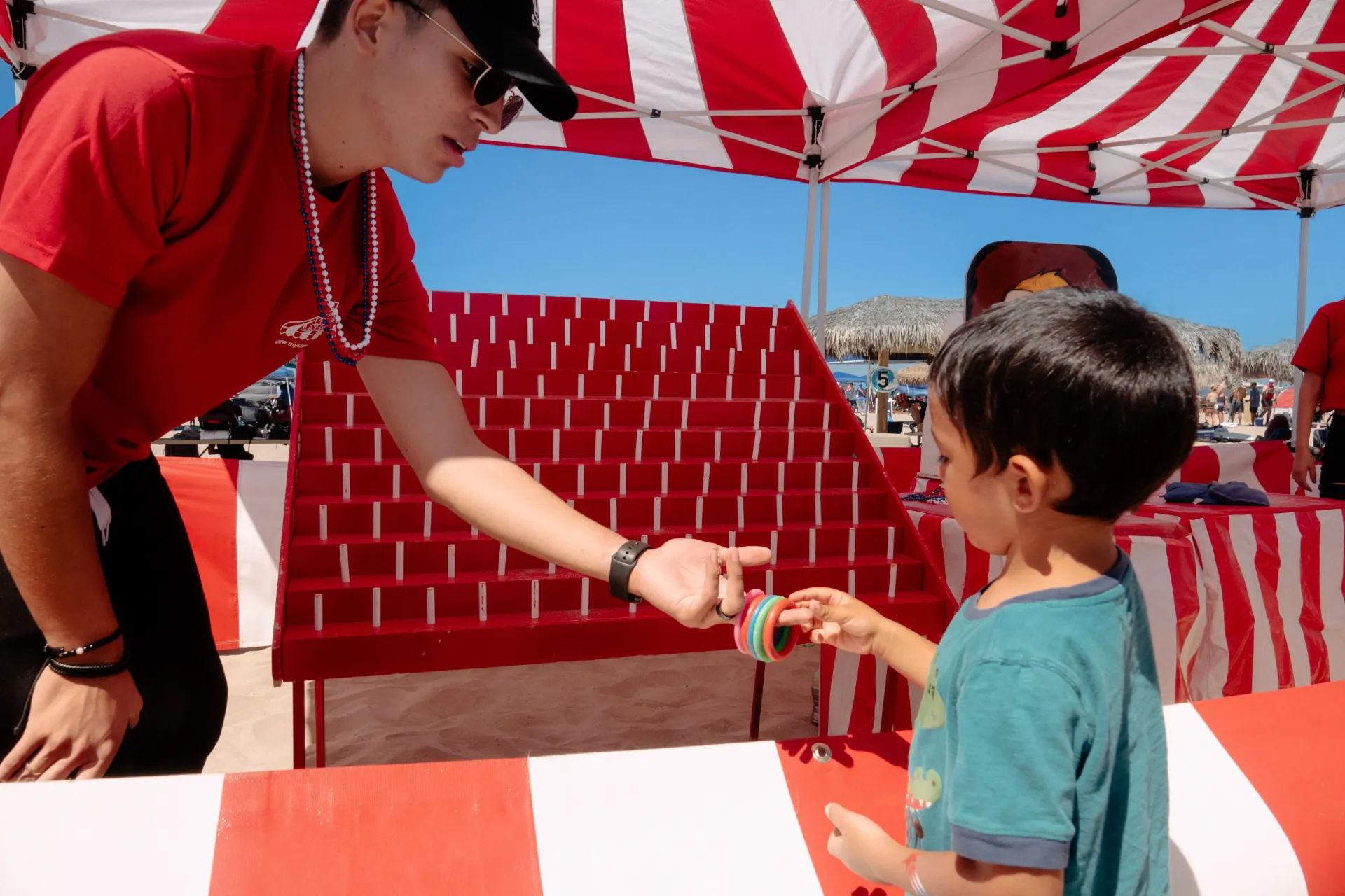 A ring toss booth on sand under a red-and-white striped canopy, with a tall red pegboard of targets and a young child reaching up to take a stack of coloured rings across the counter, blue sky and thatched beach umbrellas behind