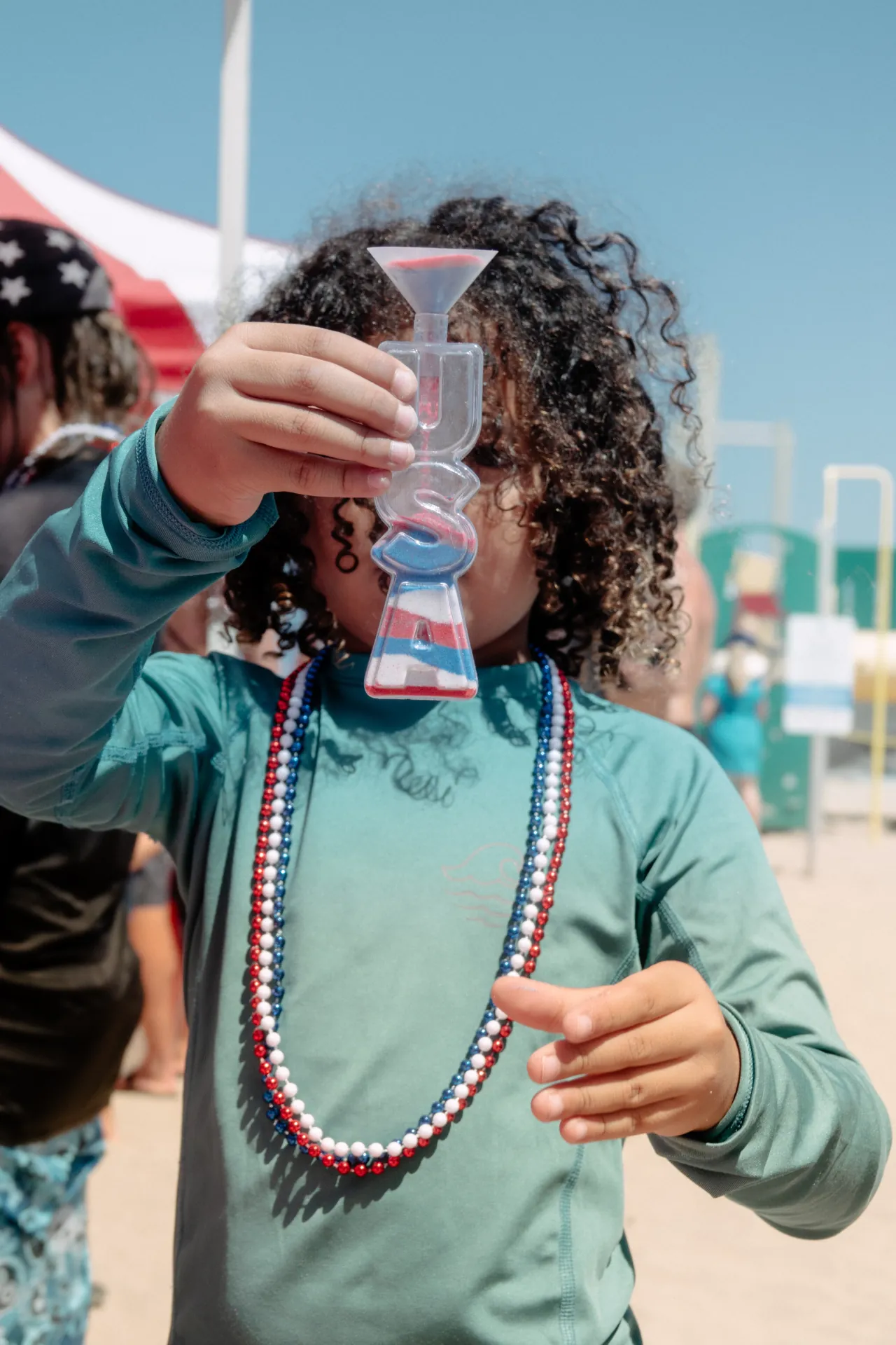A child standing on the sand holding up a USA-shaped novelty sand-art bottle layered in red, white, and blue, wearing patriotic bead necklaces, with a striped canopy and playground equipment blurred behind