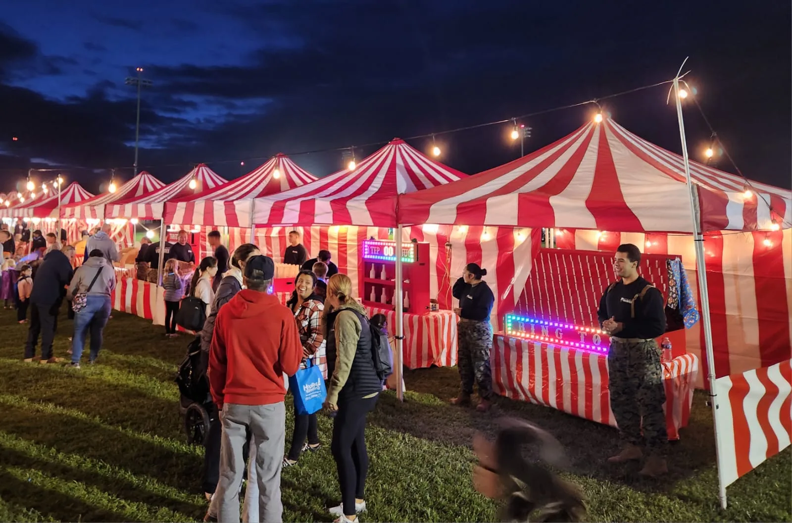 An elementary school carnival set up on a small blacktop area under oak trees, with striped booths and families walking between games in Alpine, CA